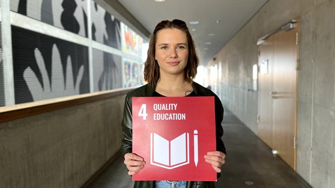Eine Frau die eine rote Karte mit der weißen Aufschrift 4 Quality Education in den Händen hält.