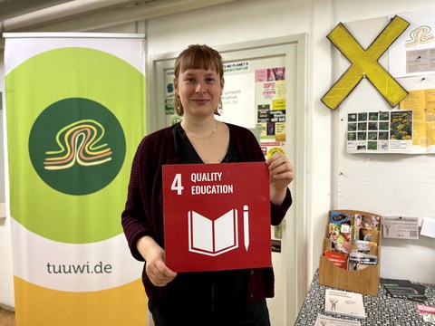 Eine Frau die eine rote Karte mit der weißen Aufschrift 4 Quality Education in den Händen hält.