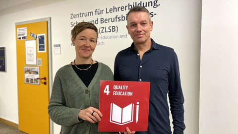 Eine Frau und ein Mann die eine rote Karte mit der weißen Aufschrift 4 Quality Education in den Händen halten.