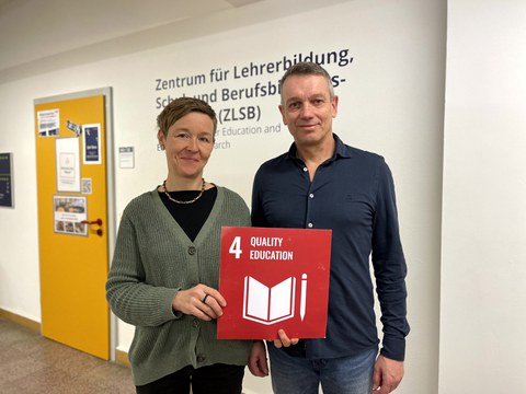 Eine Frau und ein Mann die eine rote Karte mit der weißen Aufschrift 4 Quality Education in den Händen halten.