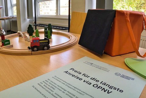 Vorn liegt ein Zettel mit dem Hinweis, dass ein Preis für die längste Anfahrt mit der Bahn vergeben wird. Im Hintergrund ist eine kleine Holzeisenbahn.