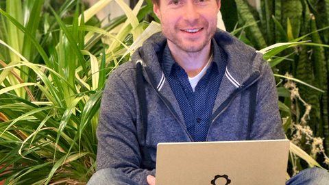 Foto von Dr. Carsten Knoll, der im Schneidersitz vor ein paar Pflanzen sitzt und in die Kamera lächelt, auf seinem Schoß liegt ein geöffneter Laptop