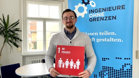 Ein Mann, der eine rote Karte mit einer 1 No poverty und einer Menschenkette in weißer Schriftfarbe in der Hand hält. Hinter ihm ist ein Plakat in hellblauer Farbe mit dem Titel Ingenieure ohne Grenzen. 