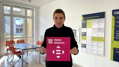 Eine Frau die eine rosafarbene Karte mit der weißen Aufschrift 10 Reduced Inequalities, darunter ein Symbol mit 4 Pfeilen und 2 parallelen Strichen in der Mitte, in der Hand hält. Im Hintergrund sieht man den Eingangsbereich vom Günther-Landgraf-Bau.