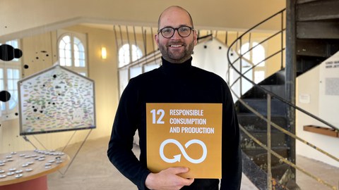Ein Mann der eine gelbe Karte mit der weißen Aufschrift 12 Responsible Consumption and Production und darunter ein unendlich Zeichen als Pfeil in den Händen hält. Im Hintergrund ist ein Foyer mit Wendeltreppe.