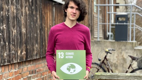 Ein Mann der eine grüne Karte mit weißer Schrift in den Händen hält mit der Aufschrift 13 Climate Action und darunter ein Symbol eines Auges mit der Landkarte drin. Im Hintergrund sieht man den Eingang der Stura Baracke. 