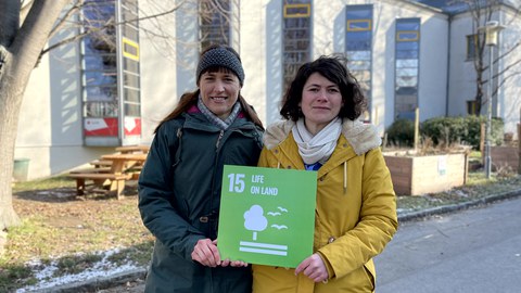 Zwei Frauen halten ein grünes Schild mit der weißen Aufschrift 15 Life on Land. darunter ein Baum mit Vögeln, in den Händen. Im Hintergrund sieht man Straße, Bäume und ein Gebäude.