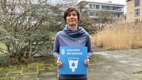 Eine Frau die ein blaus Schild mit weißer Schrift in den Händen hält, mit der Aufschrift 6 Clean Water and Sanitation und einem Symbol als Becher mit einem Wassertropfen in der Mitte. Im Hintergund sieht man Gebäude der TU Dresden und Sträucher. 