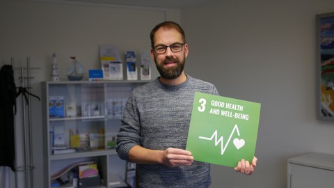 Ein Mann der eine grüne Karte mit der weißen Aufschrift 3 Good Health and Well-Being in den Händen hält. Im Hintergrund ist ein Regal mit Flyern.