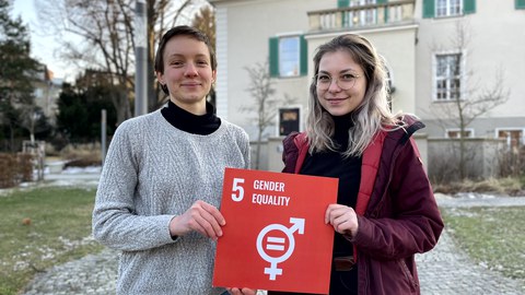 Zwei Frauen halten ein rotes Schild mit der weißen Aufschrift 5 Gender Equality, darunter eine Mischung der zwei Geschlechtersymbole in der Mitte zwei parallele Striche, in der Hand. Im Hintergrund ist ein TU Gebäude und ein geplasterter Weg.
