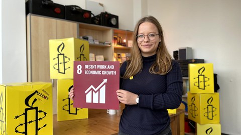 Eine Frau die eine rote Karte in den Händen hält, mit der weißen Aufschrift 8 Decent Work and Economic Growth. Darunter ein typisches Aktiensymbol in Form eines Diagramms mit aufsteigendem Pfeil. Im Hintergrund sieht man gelbe würfelförmige Kartons.