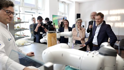 Im Vordergrund ein Mann im weißen Kittel an einer Bar, auf der rechts ein weißer Roboterarm zu sehen ist. Links davon ein hohes Glas, das mit gelben Flocken gefüllt ist, daneben eine Glasvitrine mit Speisen. Vor der Bar stehen v.l. der Sächsische Ministerpräsident, Prof. Fitzek, die Rektorin und andere Personen.