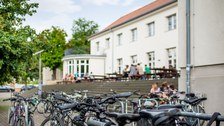 Das Foto zeigt die Treppen vor der "Alten Mensa" der TU Dresden. Oberhalb der Treppen befinden sich viele Tische, an denen einige Studierende sitzen. Im Vordergrund erkennt man viele Fahrräder. 