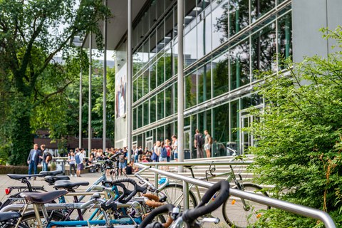 Das Foto zeigt die Außenansicht des Hörsaalzentrums der TU Dresden. Im Vordergrund stehen einige Fahrräder. Vor dem Hörsaalzentrum befinden sich viele Personen.