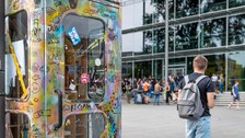 Das Foto zeigt eine bemalte Telefonzelle. Sie steht vor dem Hörsaalzentrum der TU Dresden. In der Telefonzelle befinden sich viele Bücher. Auf den Treppen vor dem Hörsaalzentrum sitzen viele Studierende. 
