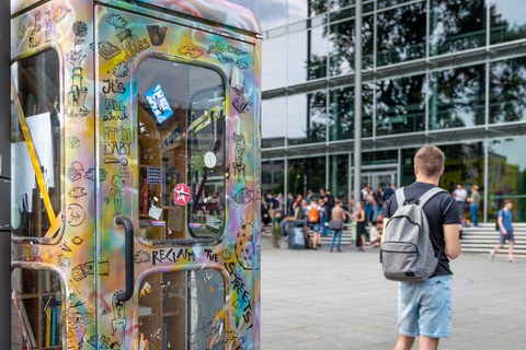 Das Foto zeigt eine bemalte Telefonzelle. Sie steht vor dem Hörsaalzentrum der TU Dresden. In der Telefonzelle befinden sich viele Bücher. Auf den Treppen vor dem Hörsaalzentrum sitzen viele Studierende. 