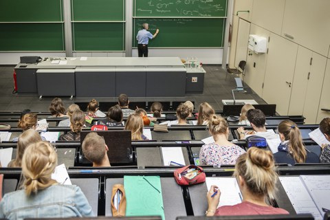 Foto von oben: Ein Dozent schreibt an der Tafel in einem Hörsaal mit aufmerksamen Studenten.