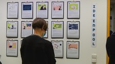 An einer Wand hängen mehrere Klemmbretter mit A4 Seiten. Eine Frau steht davor und liest sich den Text darauf durch.