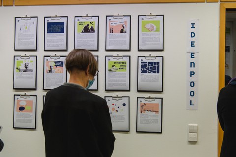 An einer Wand hängen mehrere Klemmbretter mit A4 Seiten. Eine Frau steht davor und liest sich den Text darauf durch.