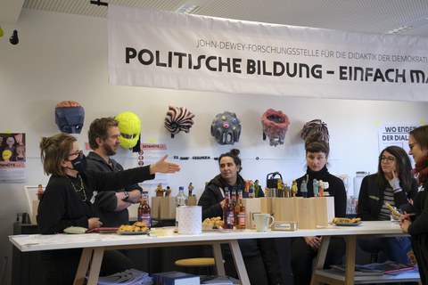 Mehrere Personen sitzen um einen Tisch. Darauf befinden sich Essen, Getränke, und Modellfiguren. Im Hintergrund hängen Masken und ein Banner auf dem "Politische Bildung" steht.