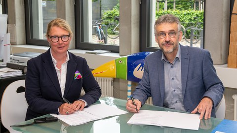 Links eine Frau, rechts ein Mann an einem Schreibtisch. Vor sich aufgeschlagene Mappen.