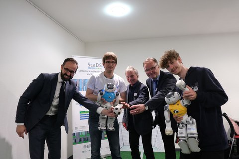 Die fünf genannten Herren stehen nebeneinander, etwas vorgebeugt, und strecken jeweils eine Hand aus.  Hr. Gemkow hält einen kleinen Fußball in der Hand,  Hr. Jagla und Hr. Loos halten jeweils einen kleinen Roboter.