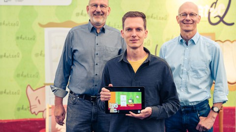 Drei Herren schauen in die Kamera, der junge Mann in der Mitte hält ein Tablett in der Hand.