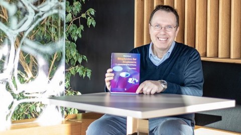 Prof. Schießel sitzt an einem Tisch und hält sein Buch in die Kamera.