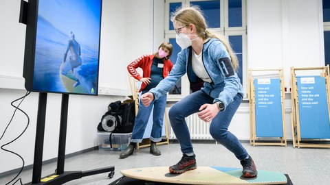 Ein junges Mädchen steht in Surferhaltung auf einem Podest vor einem Bildschirm. Im Hintergrund beobachtet eine Frau das Geschehen.