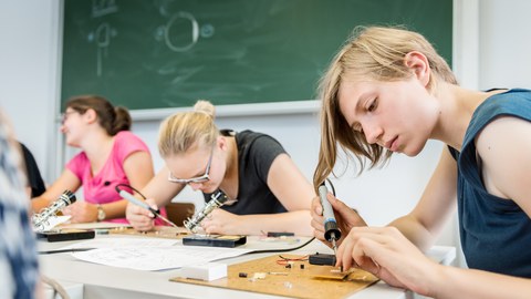 Drei junge Frauen sitzen an Tischen vor einer Tafel und arbeiten mit Lötkolben.
