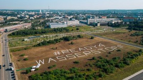 Landschaft, im Hintergrund Industrie. Auf einem Feld die Botschaft "Ready 4 Wasserstoff"
