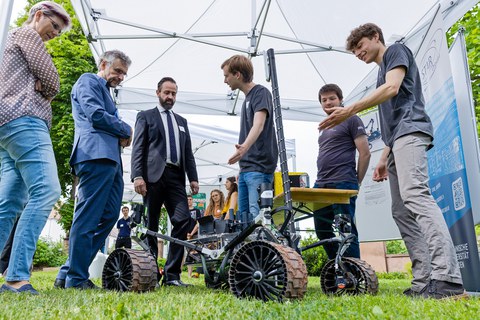 Fotoaufnahme von mehreren Personen, die sich im Freien unter dem Dach eines Zeltes befinden. Sie sind in einer Unterhaltung. Am unteren Bildrand befindet sich ein kleines, sehr technisch wirkendes, Fahrzeug.