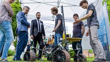 Fotoaufnahme von mehreren Personen, die sich im Freien unter dem Dach eines Zeltes befinden. Sie sind in einer Unterhaltung. Am unteren Bildrand befindet sich ein kleines, sehr technisch wirkendes, Fahrzeug.