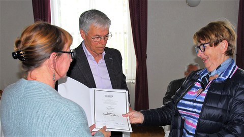 Drei Personen stehen im Kreis, es wird die Goldene Diplomurkunde überreicht.
