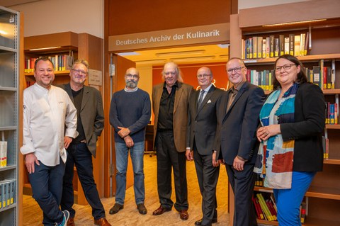 6 Männer und eine Frau stehen in einer Bibliothek um einen Durchgang herum. Über dem Durchgang der Schriftzug "Deutsches Archiv der Kulinarik"