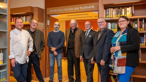 6 Männer und eine Frau stehen in einer Bibliothek um einen Durchgang herum. Über dem Durchgang der Schriftzug "Deutsches Archiv der Kulinarik"
