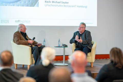 Zwei Herren, in Sesseln auf einem Podium sitzend, im Gespräch. 