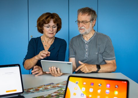Ein älterer Herr hält ein Tablett in der Hand, eine ältere Dame sitzt neben ihm. Beide schauen auf das Tablett