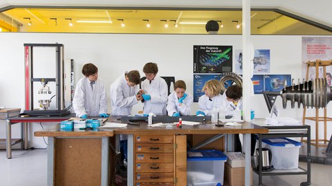 Eine Gruppe von Schüler:innen in weißen Laborkitteln schaut auf einen Tisch