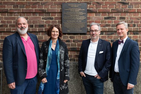 Gruppenfoto (eine Frau, drei Männer) vor einer Ziegelwand. Daran eine Gedenktafel "Historische Stätte der Chemie"