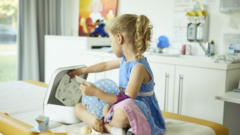 Kleines Mädchen sitzt vor einem geöffneten Kinderkoffer auf einem Krankenhausbett