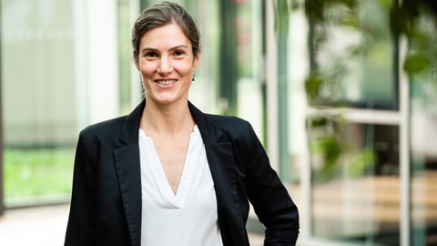 Portrait einer dunkelhaarigen Frau mit weißer Bluse und schwarzem Blazer vor grünem Hintergrund.