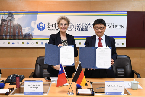 Eine Frau und ein Mann stehen hinter einem Tisch mit deutschen und taiwanesischen Flaggen. Sie halten Urkunden in die Kamera.