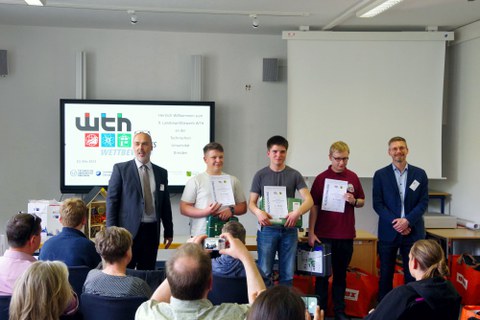 Gruppenfoto mit den 3 Siegern des Landeswettbewerbs WTH, links und rechts davon je ein Herr im Anzug. Davor sitzen Menschen auf Stühlen und schauen zu den Preisträgern.