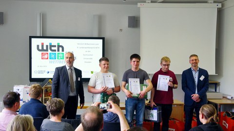 Gruppenfoto mit den 3 Siegern des Landeswettbewerbs WTH, links und rechts davon je ein Herr im Anzug. Davor sitzen Menschen auf Stühlen und schauen zu den Preisträgern.