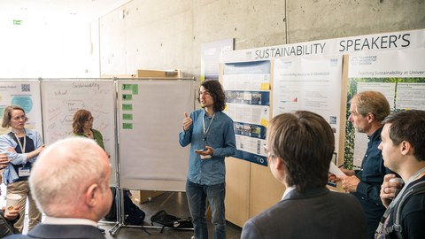 Fotoaufnahme einer Person, die vor Flipcharts und plakatierten Stellwänden steht und zu einer Gruppe an Personen spricht.