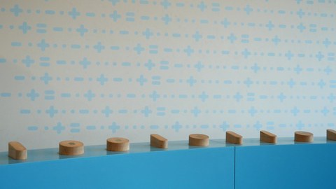 Blauer Sockel mit Holzzylindern in verschiednen Größen vor heller Wand. 
