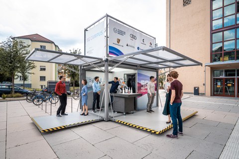Mehrere Personen stehen in einem mobilen Präsentationsstand, der die Überschrift "5G Campus" und die Logos der Sponsoren trägt.