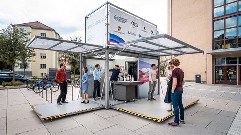 Mehrere Personen stehen in einem mobilen Präsentationsstand, der die Überschrift "5G Campus" und die Logos der Sponsoren trägt.