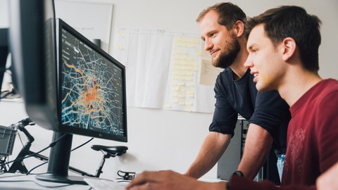 Fotoaufnahme von zwei Männern, die auf einen Computer-Bildschirm schauen. Dieser zeigt eine Karte von Dresden, auf der Fahrradrouten farbig markiert sind.